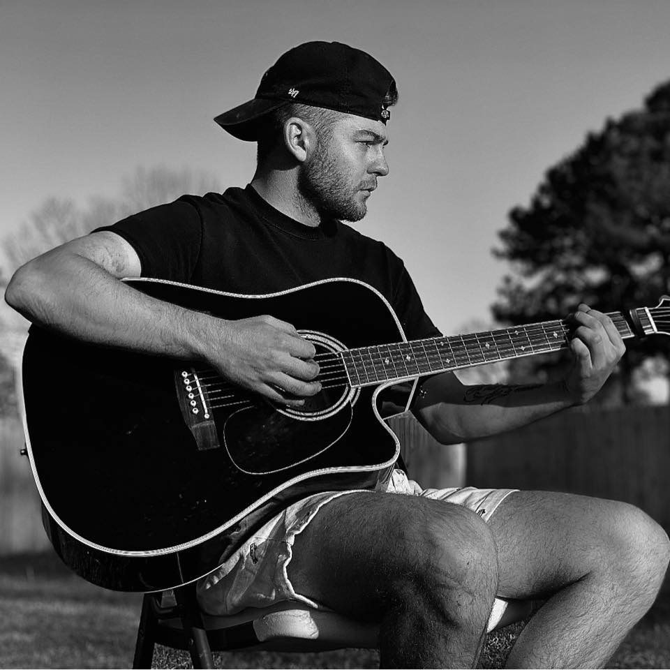 Robbie Bradshaw playing guitar with ball cap on backwards and wearting shorts