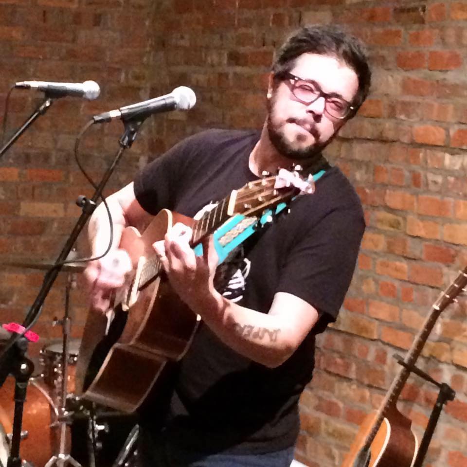 Derek Smith playing guitar. Brick wall background.