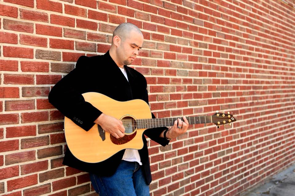 Man stanind in front of brick wall playing guitar