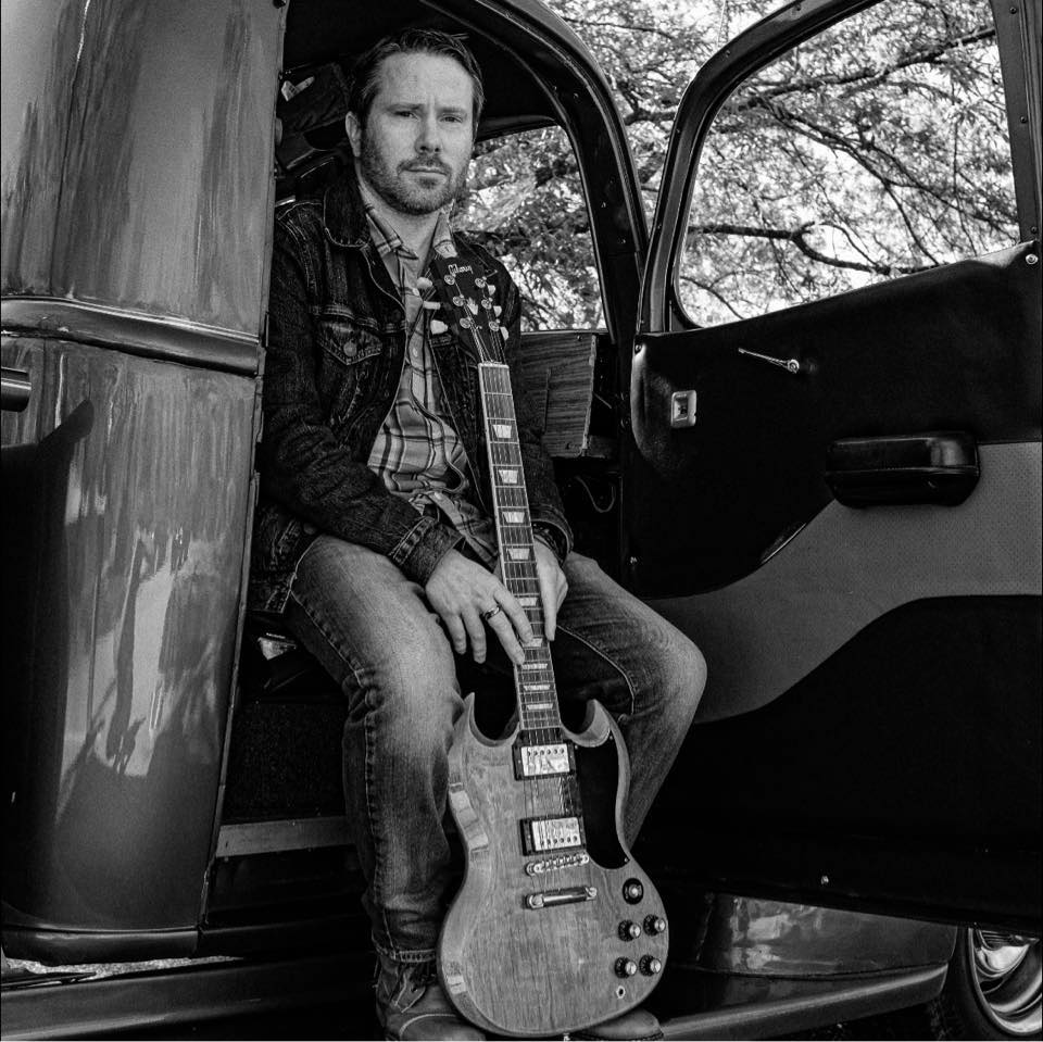 Rob Oliver sitting in an old truck holding a guitar