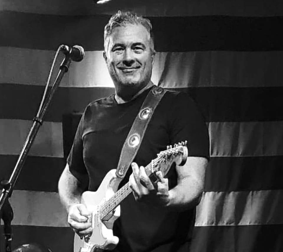 Bryan Dunn holding guitar stading in front of microphone with US flag backdrop